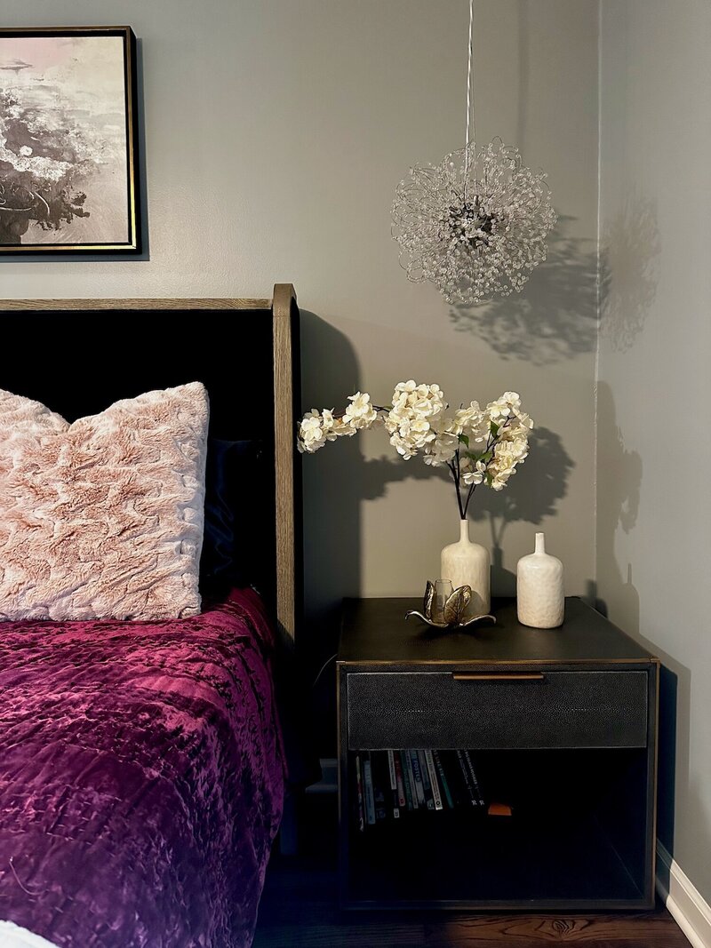Bed with purple velvety sheets and textured pillows next to a night stand