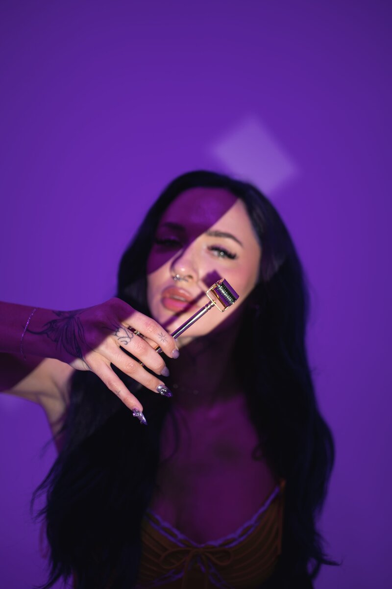 Woman with dark, long hair and tattoos on her arm side-eyes the camera with a spotlight on her face and moody purple background as she holds a small metal roller in front of her cheek.