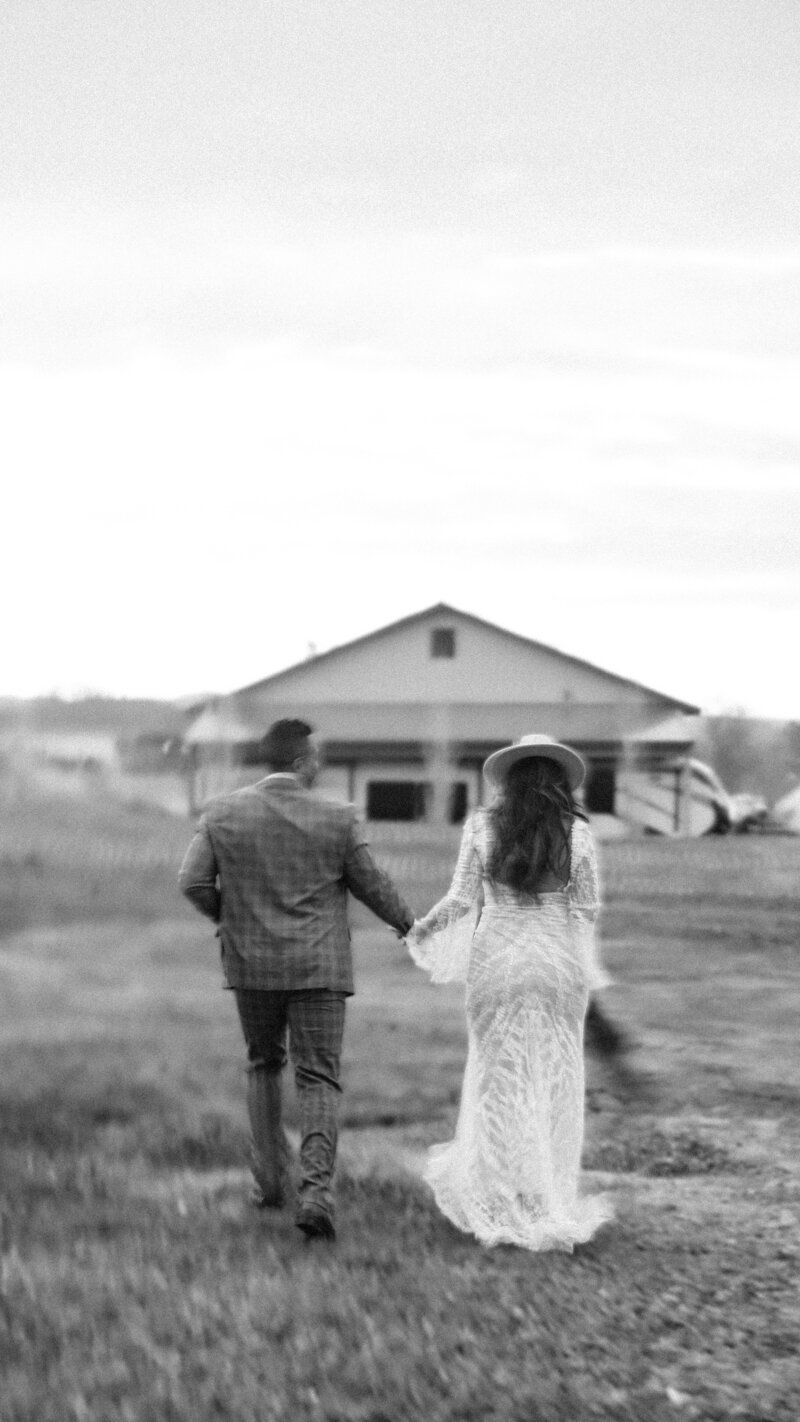 Couple in black and white walking away from camera toward a barn.