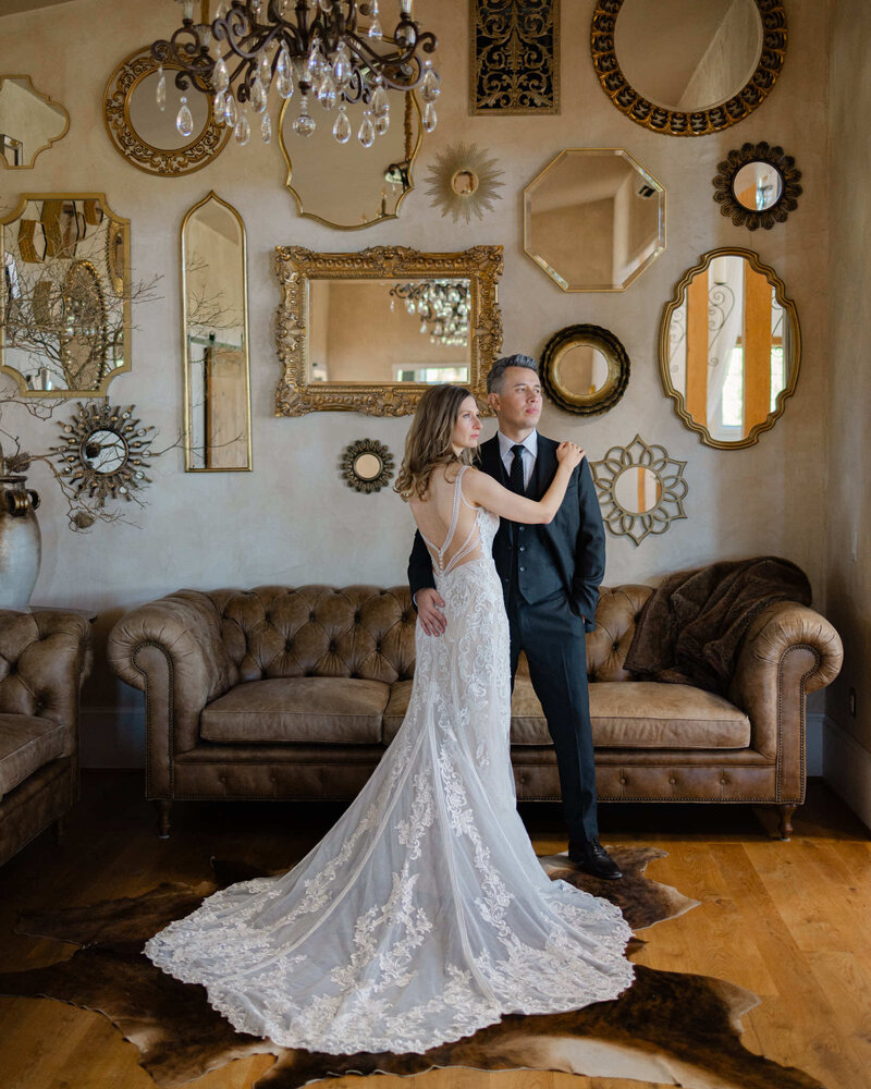 Couple posing in their wedding attire in a library with mirrors and sofa in the background.
