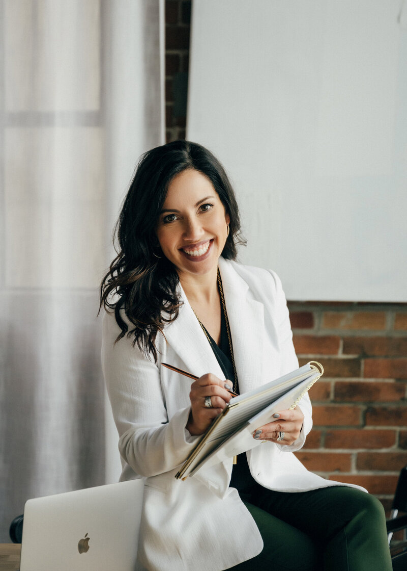 Jessica wearing a white blazer, smiling and writing in a spiral bound planner