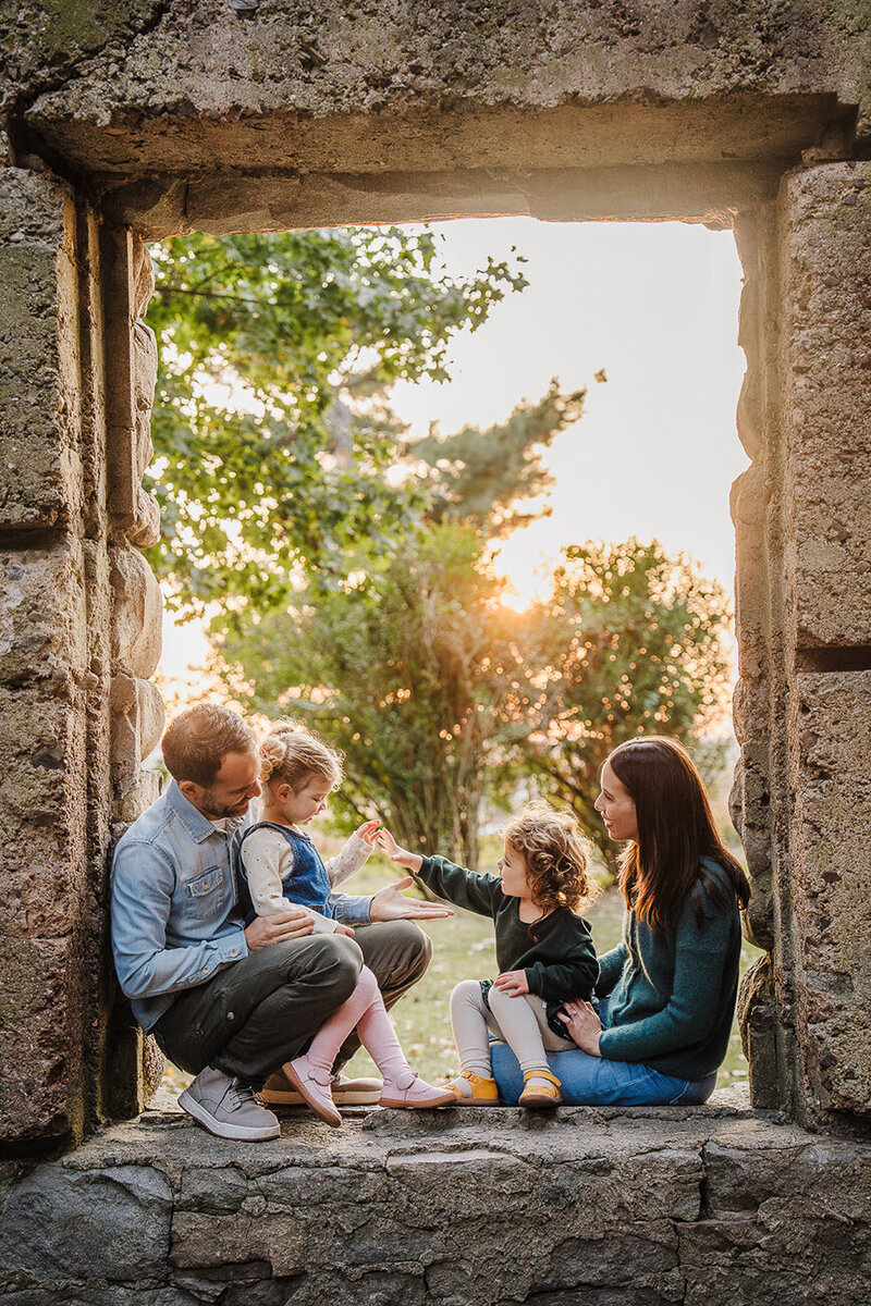 family with twin toddler daughters sit in the window of a castle ruin outside boston