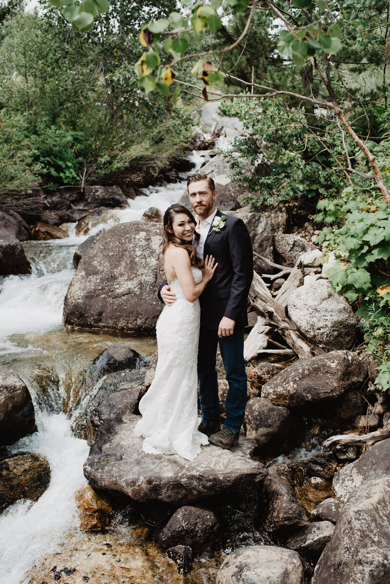 wyoming elopement pictures with bride adn groom standing on a rock in front of a waterfall in the woods of the grand tetons captured by jackson hole photographers