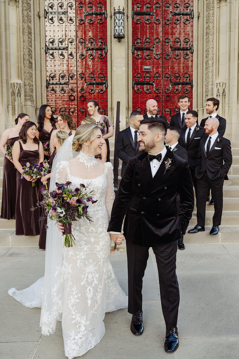 Bride and groom exiting a historic church with vibrant red doors, surrounded by their wedding party dressed in elegant deep-toned attire.