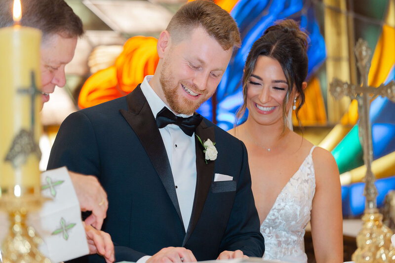 a smiling bride and groom sign the church register during their wedding ceremony.  Captured by Ottawa wedding photographer JEMMAN Photography