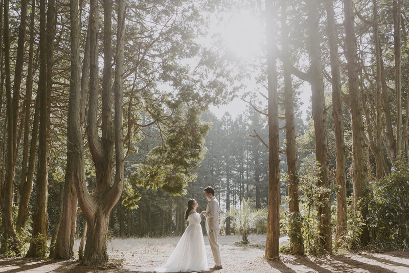 bride in white dress and groom in tan suit hold hands in Secret Forest in Jeju Island