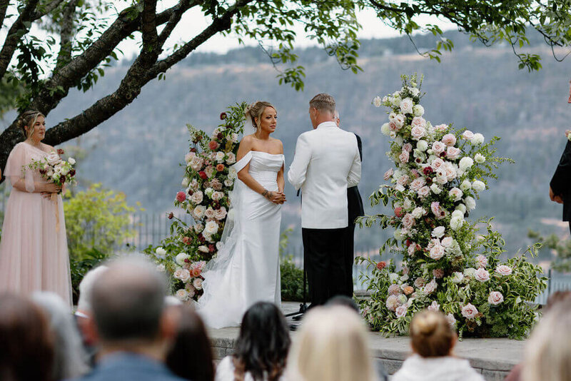 A candid wedding ceremony photo taken from behind the guest seats. The couple is standing on a stone stage facing each other. The bride is on the left wearing an all white wedding dress that has off-the-shoulder sleeves. The groom is on the right and is wearing a white and black tuxedo. The couple is standing between a lush, freestanding floral arch that is broken into two sections. These are like floral pillars. The flowers are a soft blend of white, cream, blush, pink, and burgundy wedding flowers. The background scenery is an out of focus view of the Columbia River Gorge. The location is  the Columbia Gorge Hotel and Spa in Hood River, Oregon.
