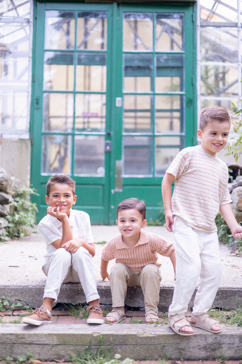Three young brothers smile and play on stone steps in front of vibrant green glass doors during a lighthearted outdoor family session in Greenwich, CT. Captured by Nicole Neira Photography, this joyful moment highlights the boys' personalities and sibling bond.