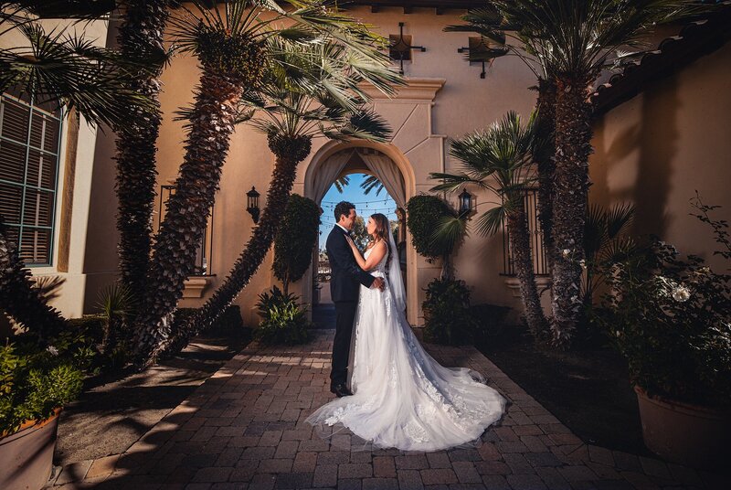 A bride and groom embrace in front of Talega Golf Club's main entry archway, displaying Spanish architecture influence, on their wedding day.