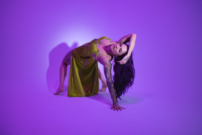 Woman with long, black hair is in a backbend with one arm reaching over her head as she stares at the camera wearing a silky green dress in front of a purple backdrop.