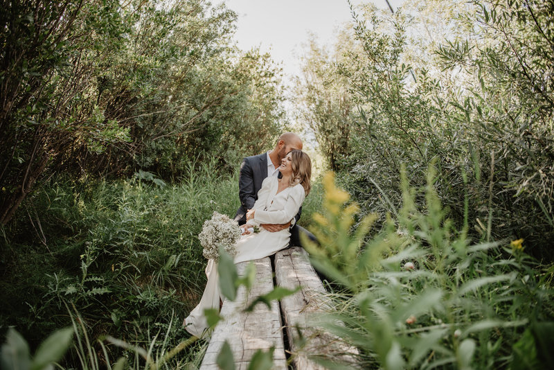jackson hole wedding photographers captures groom holding his bride as they sit on a fallen log together in a pond as the bride laughs and leans into her groom
