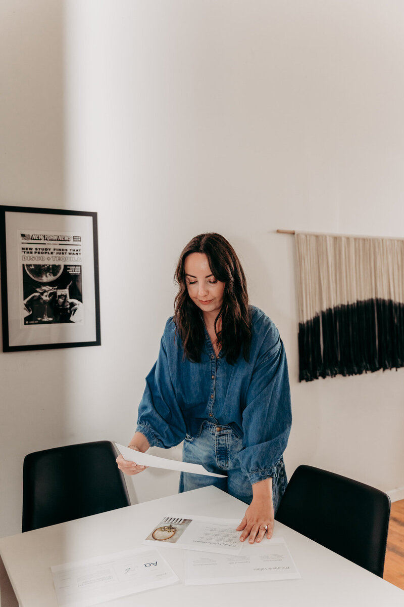 Delia, copywriter, holding up a brand strategy deck by a table, with a denim top and pants.