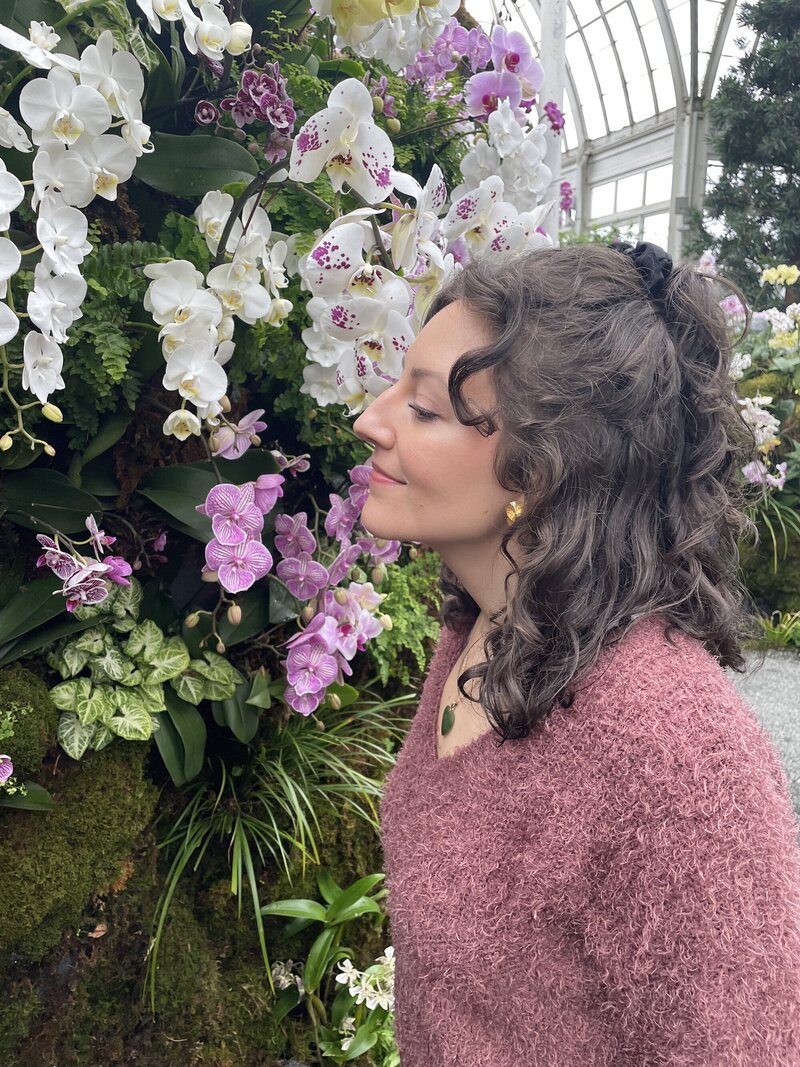 A mindfulness mentor with brown, short, curly hair stands in a lush botanical garden, surrounded by beautiful pink and white flowers. She stops to smell the flowers, taking a moment to practice mindfulness and connect with nature. The image evokes feelings of peace, serenity, and mindfulness, making it perfect for anyone interested in meditation and self-care.  [Nicole QW]