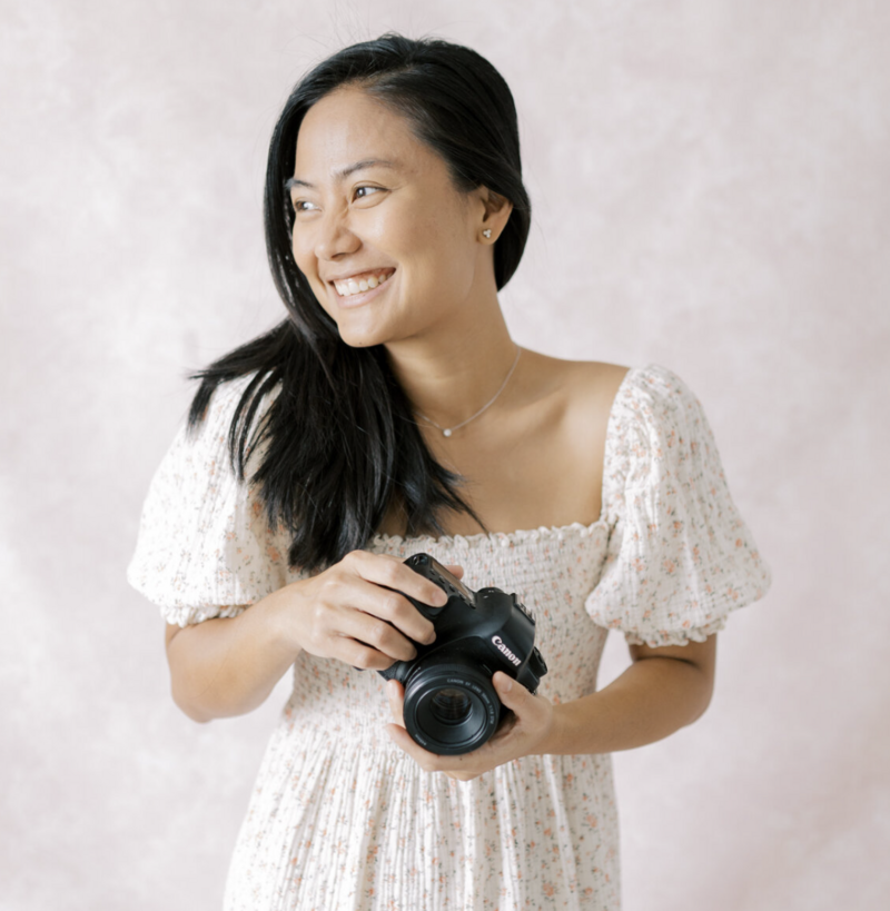 Image of Photographer Ericka Ana looking away, holding her camera and smiling. She is wearing a beautiful white dress with small floral details.