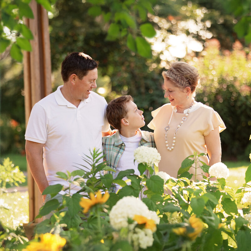 Family standing together in a sunny garden, with two adults and one child smiling at each other among blooming white flowers and greenery.