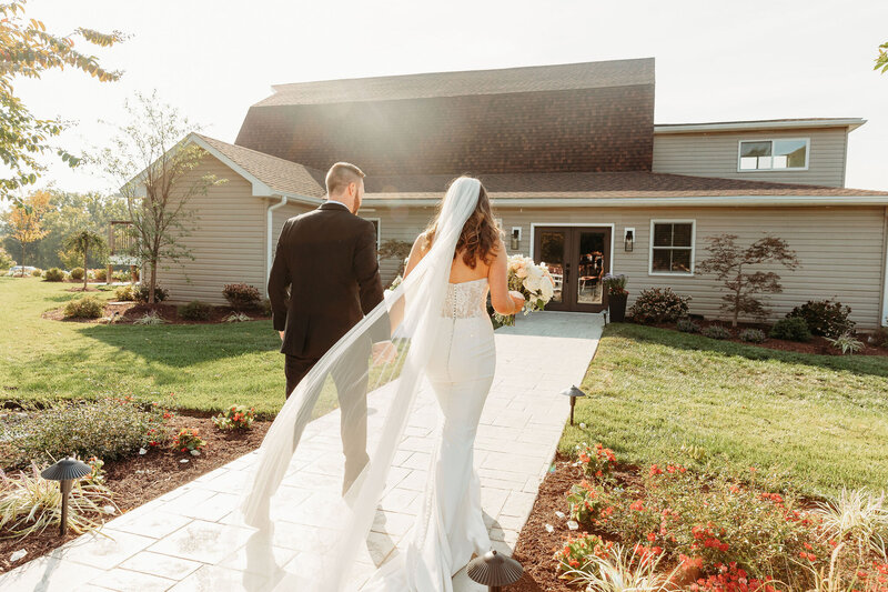 A bride and groom walk hand in hand on a sunlit path leading to a charming house. The bride wears a flowing white dress and veil, while the groom is in a black suit. Set amidst lush greenery and vibrant flowers, the scene evokes the charm of elegant reception venue galleries.
