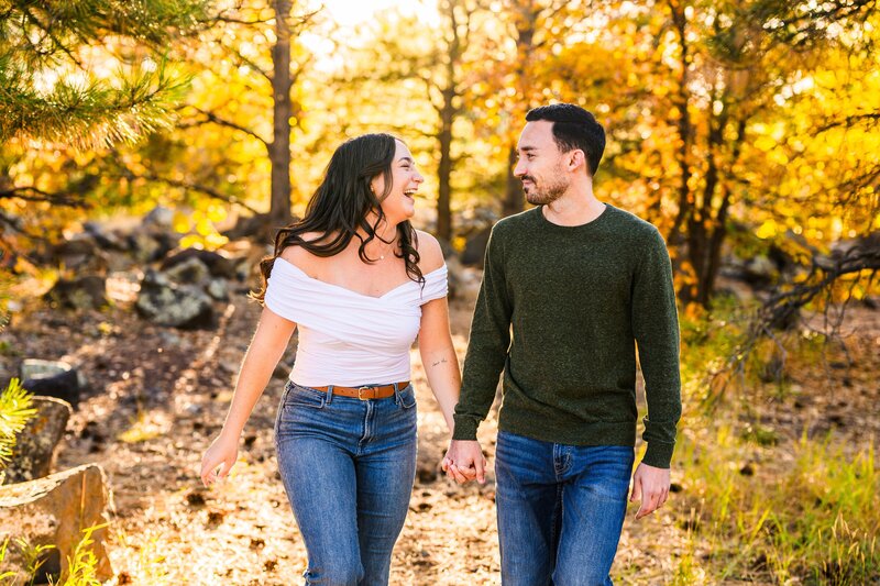 Engaged couple holding hands and walking towards camera looking at each other smiling and laughing with orange fall leaves behind in autumn season in Flagstaff Arizona