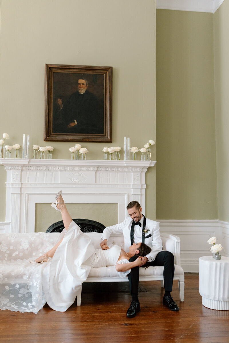 A playful and relaxed wedding photo of a bride lying across a couch with her groom beside her, in a vintage styled room.