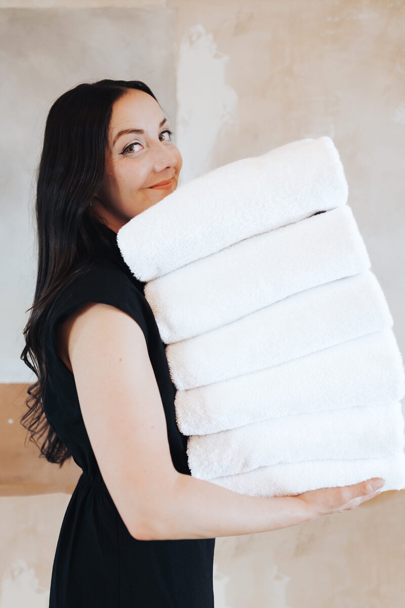 Woman with long, dark hair wears a black dress while smiling to the camera and holding a stack of 6 white, folded towels.