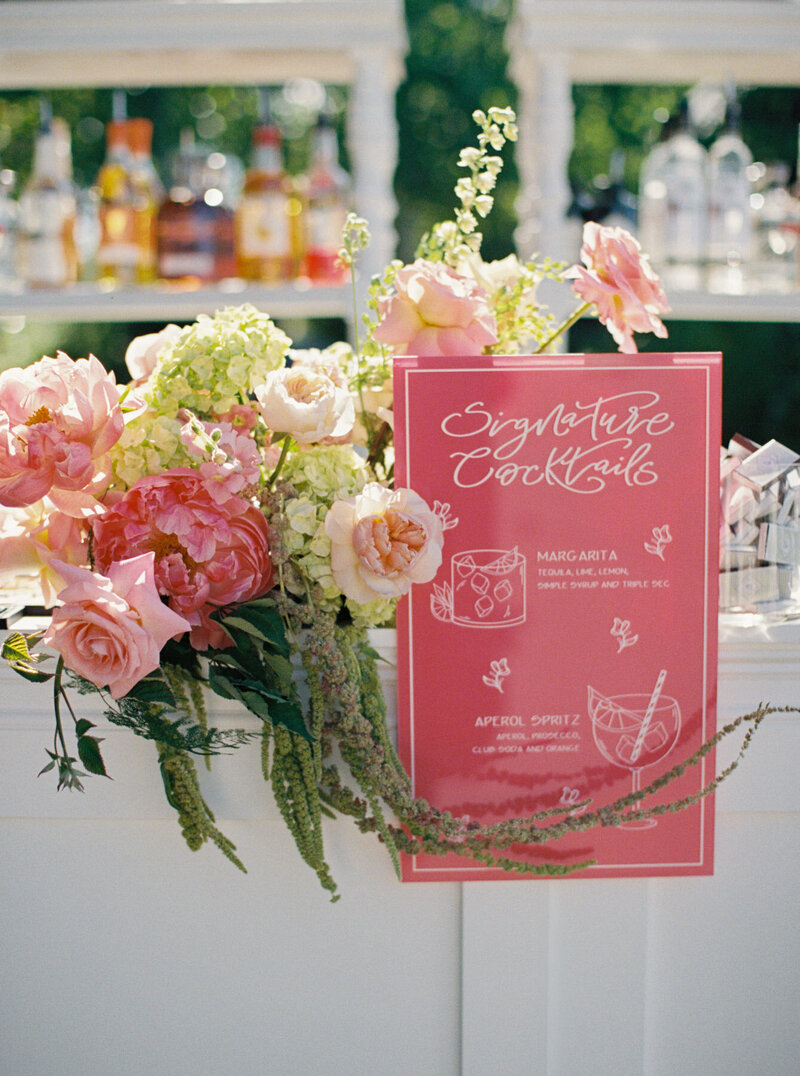 Signature cocktails sign with customized calligraphy sits on a bar top next to flowers at a wedding.

