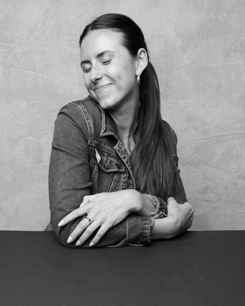 Black and white portrait of a woman sitting at a table with her arms gently wrapped around herself, eyes closed and smiling with contentment. She wears a denim jacket, hoop earrings, and a ring, exuding warmth and self-assurance. A serene, unfiltered moment from 'THIS IS ME – A Photography Experience by Katie Campbell' in Atlanta, GA, celebrating self-love and authenticity.