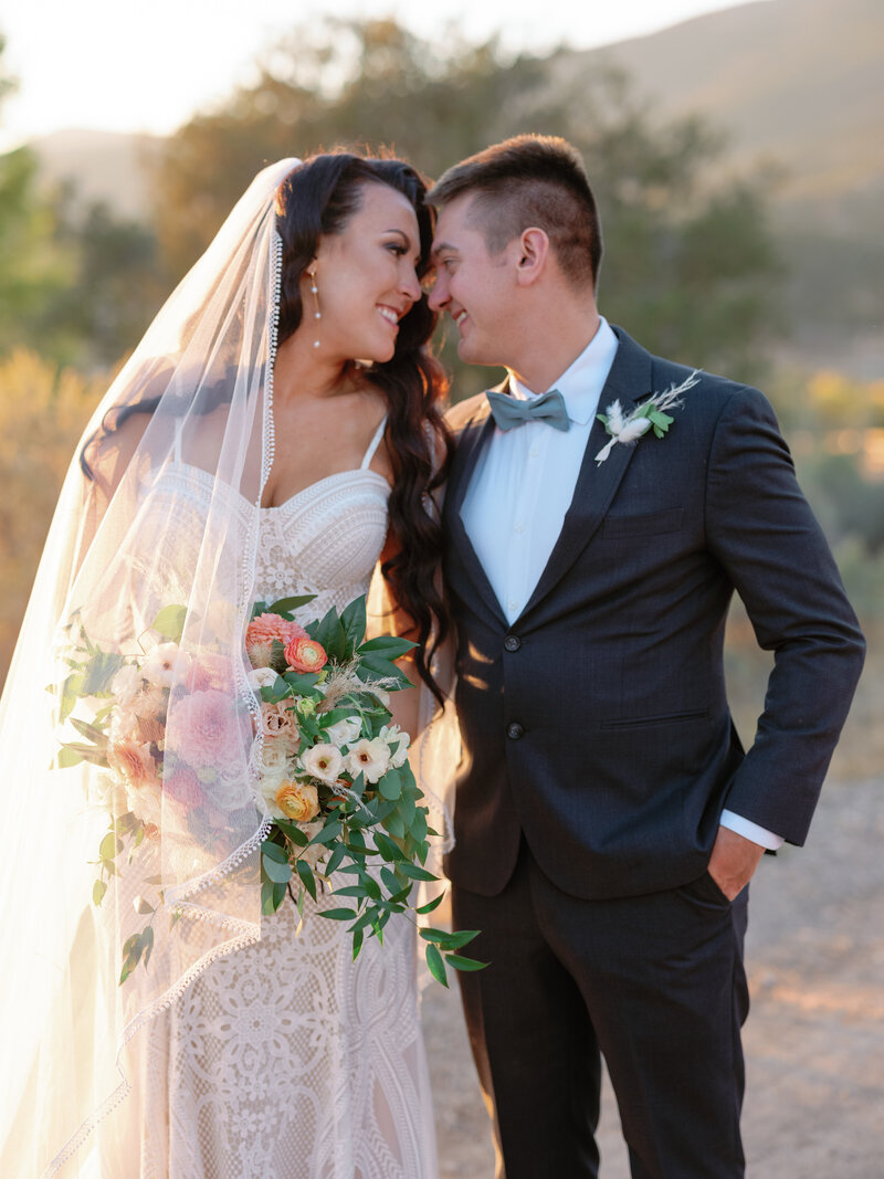 Bride and groom holding large bouquet, with a long lace trimmed veil sweeping over.