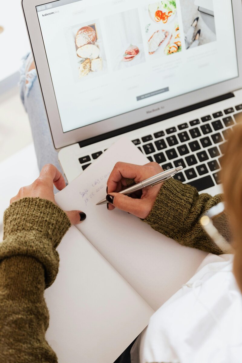 A woman writes on a notepad. Her hands, the notebook, her Macbook laptop are shown in top down view