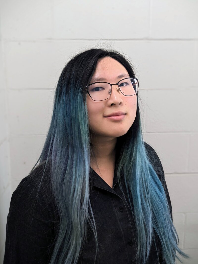 A person with long, straight hair dyed in shades of blue and black, wearing glasses and a black button-up shirt. They are standing against a light-colored cinder block wall, looking directly at the camera with a neutral and confident expression.