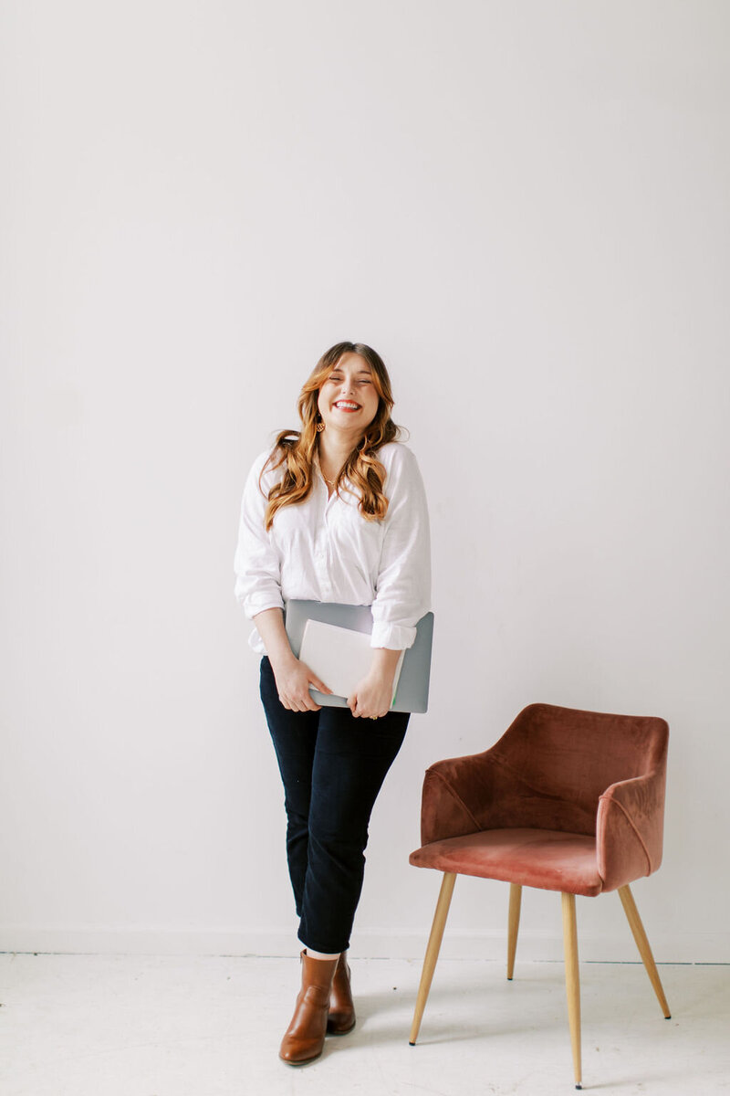 Amber is standing beside a pink velvet chair and wearing a long sleeve white button up with black pants and brown boots. She is holding her laptop and a book while smiling at the camera.