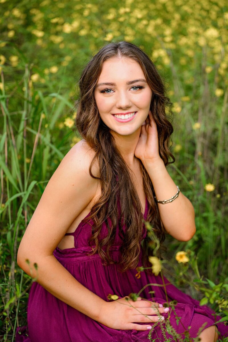 portrait of a senior girl with her hand by her face in a field of yellow flowers wearing a purple dress