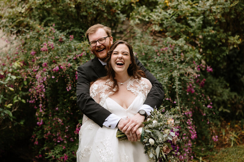 A bride in a flowy white gown holds a lush bouquet while embracing her partner in front of vibrant purple and green foliage.