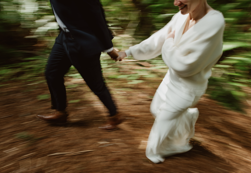 A couple runs hand-in-hand down a trail in Redwood National Park during their all-day intimate redwood elopement