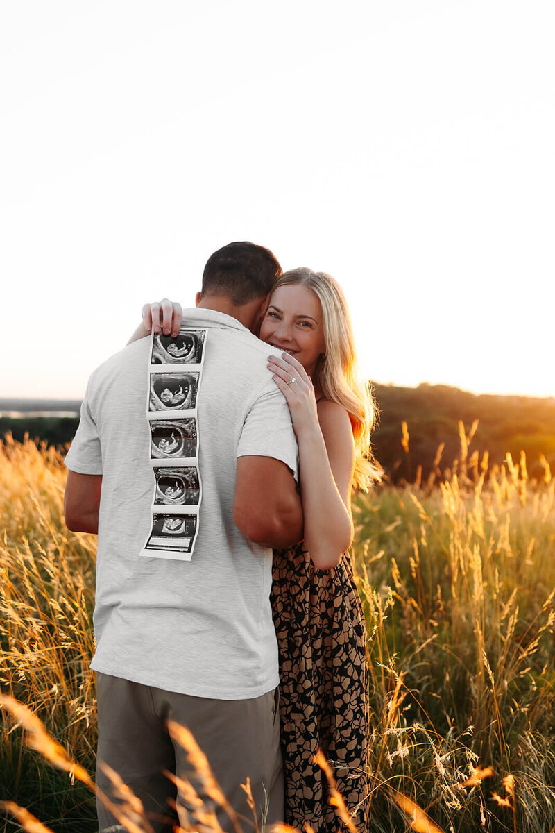 Expecting mother smiles holding photos of her baby while her husband hugs her