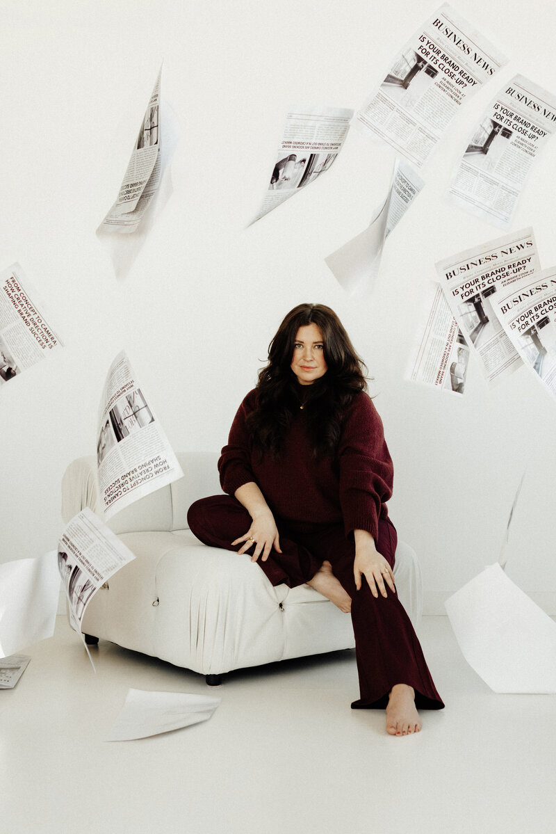 Creative portrait of Elisabeth Eden sitting on a soft white chair with falling newspapers around her, showcasing a modern and artistic brand photography concept.