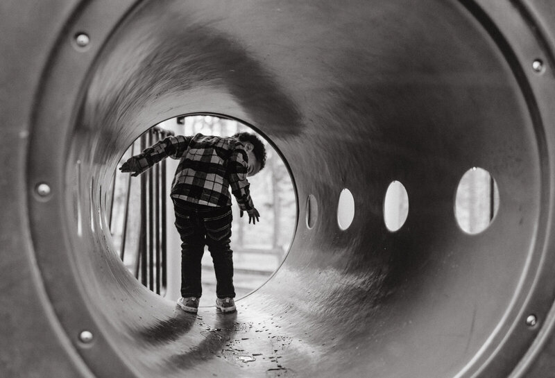 Black and white portrait of a little boy exploring a playground tunnel. 