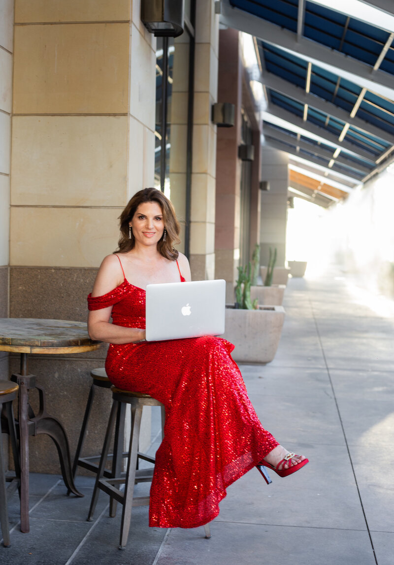 Melanie is in a red gown with red heels, seated on a stool with her laptop on her lap and open