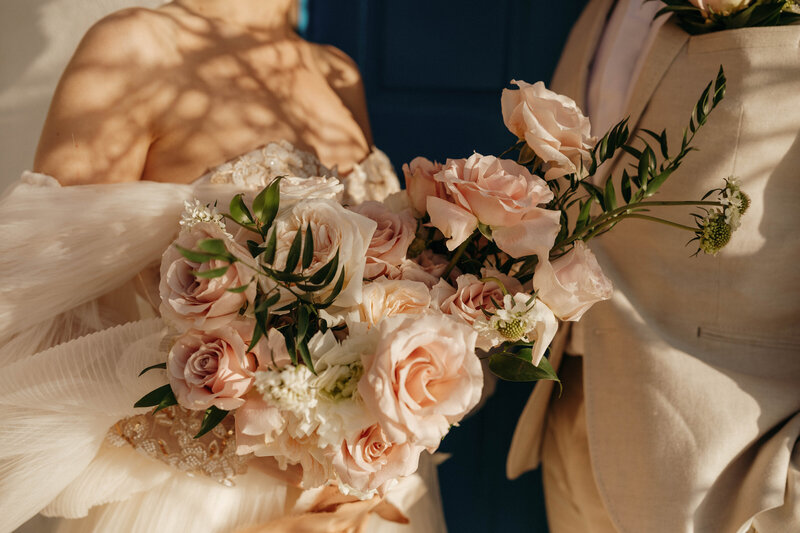 Close-up of a sunlit blush and cream rose bridal bouquet with greenery, held between a bride and groom in warm, golden afternoon light.