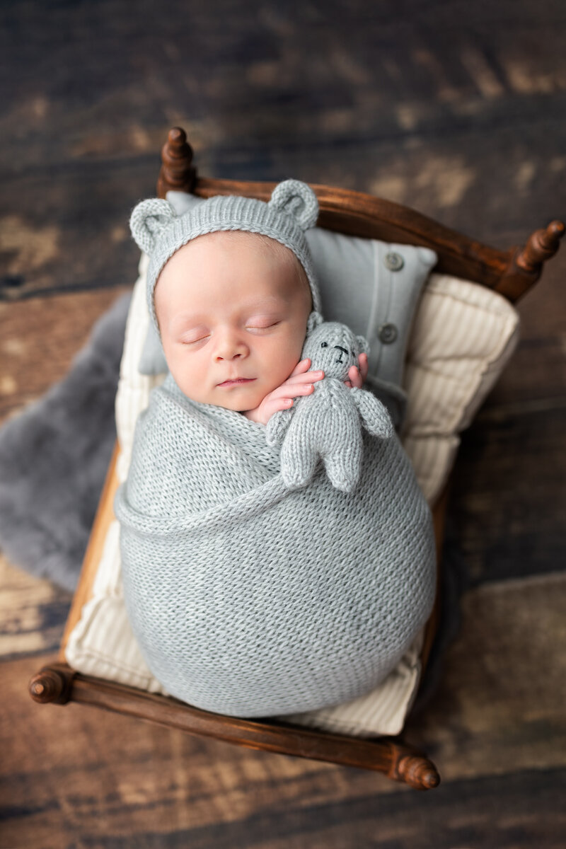 Soft blue knits and a tiny teddy—this newborn boy rests in pure comfort on a handcrafted baby bed.