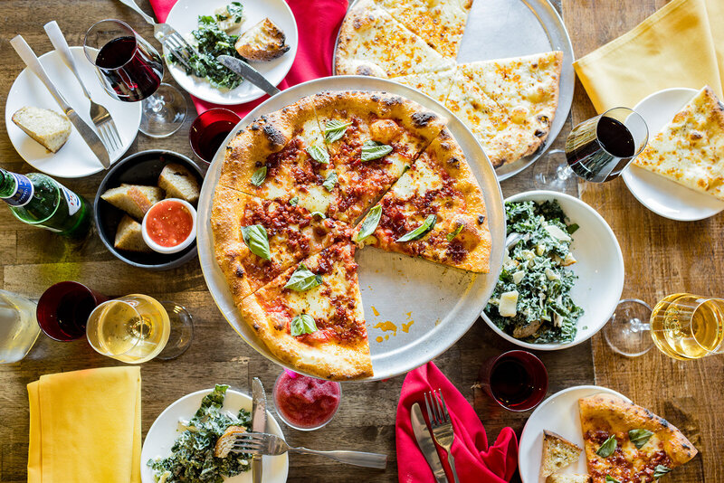 A vibrant flat-lay of an Italian dining experience featuring artisan pizzas, fresh kale Caesar salad, crusty bread with marinara, and an assortment of red and white wines. Captured in a Columbia, SC, restaurant, this commercial food photography showcases a warm and inviting atmosphere, perfect for branding local eateries.