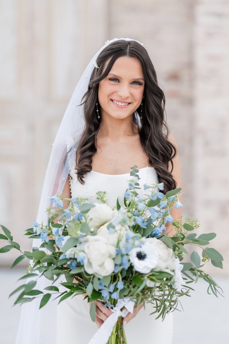 A bride stands while smiling with her large white and blue bouquet