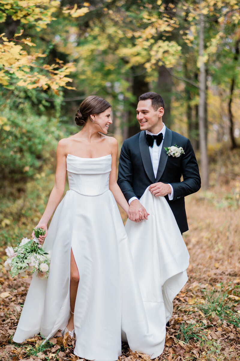 Groom holds his brides wedding skirt as they walk through the autumn leaves