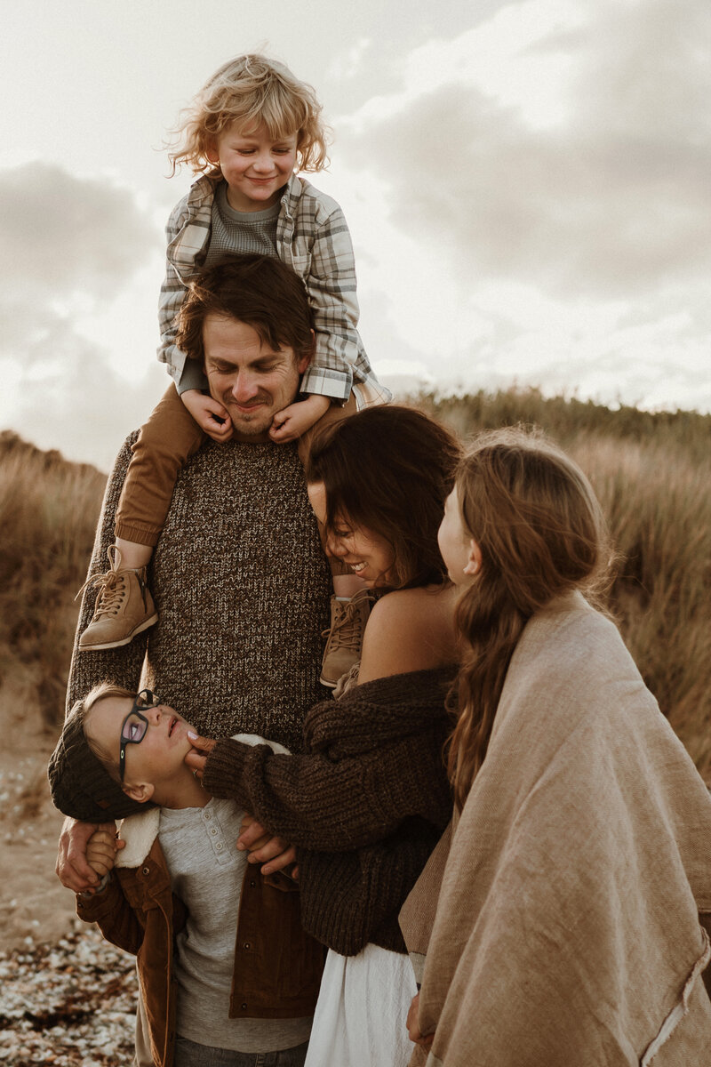 Family photography, family of five at a beach location. Dad has one son on his shoulders while mother embraces her daughter and touches her younger sons face as he looks up to her. Family are all close and connected in the image