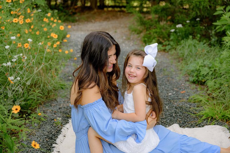 daughter dressed in white with a white hair bow laughs as her mother whispers in her hear