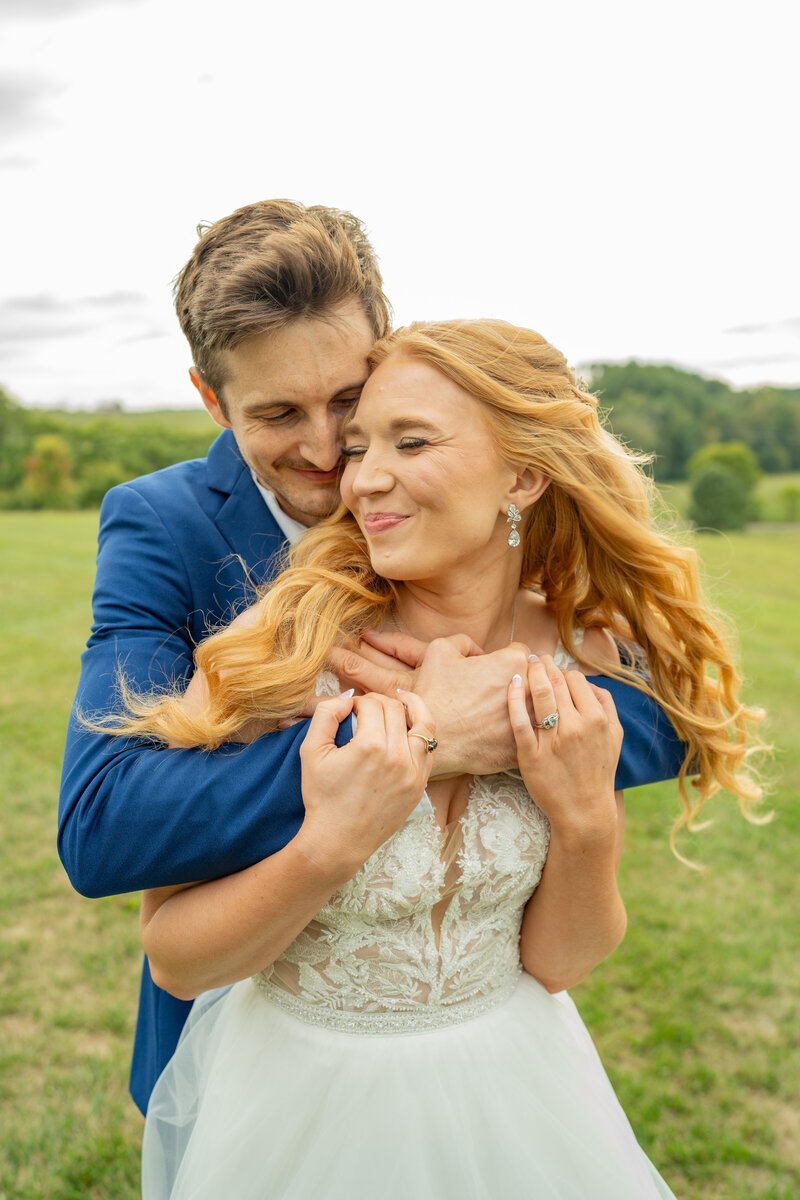 The groom is hugging the bride from behind with mountains in the background