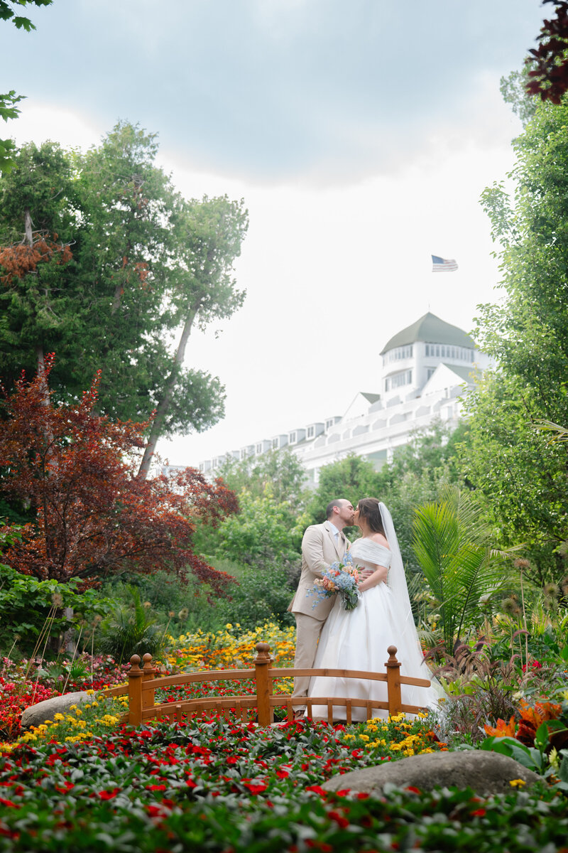 A bride and groom kiss on a little bridge in front of the Grand Hotel in the flowering secret garden on Mackinac Island
