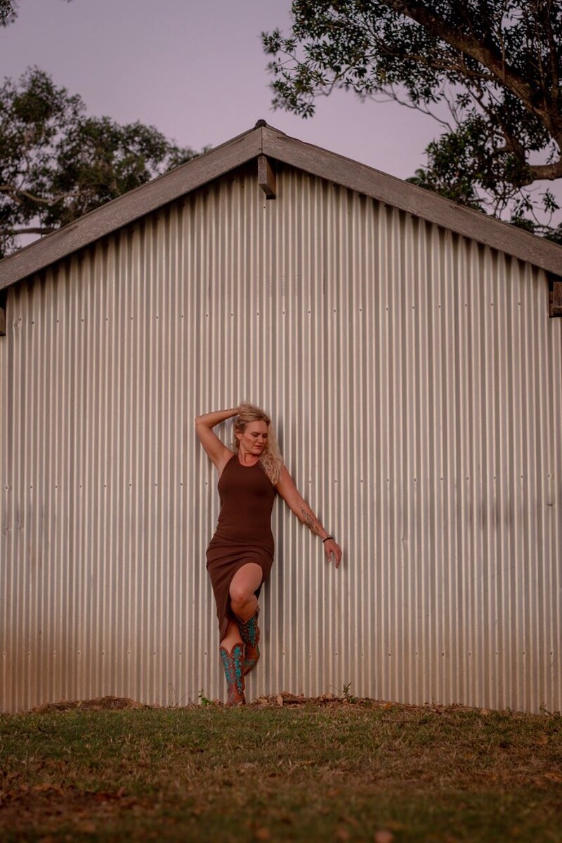 woman in embodied pose leaning on barn in red dress.