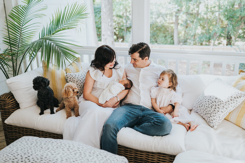 Family of four with two mini poodles sits on white outdoor sofa looking at newborn baby boy during photo session