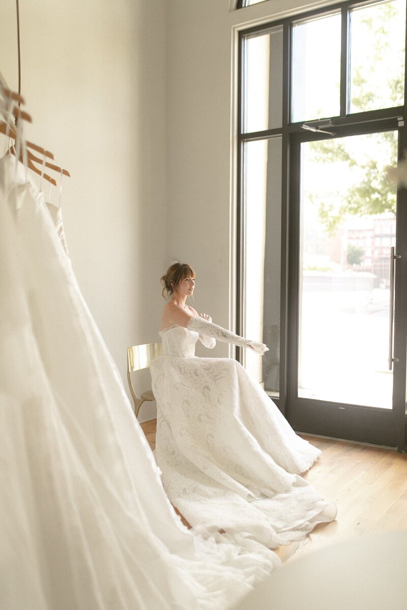 Bride in an off-shoulder lace gown sitting by a large window.