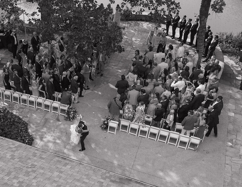 Aerial view of a wedding ceremony in black and white. Guests are seated outdoors, forming a semi-circle. A couple dances in the foreground, creating a joyful atmosphere.