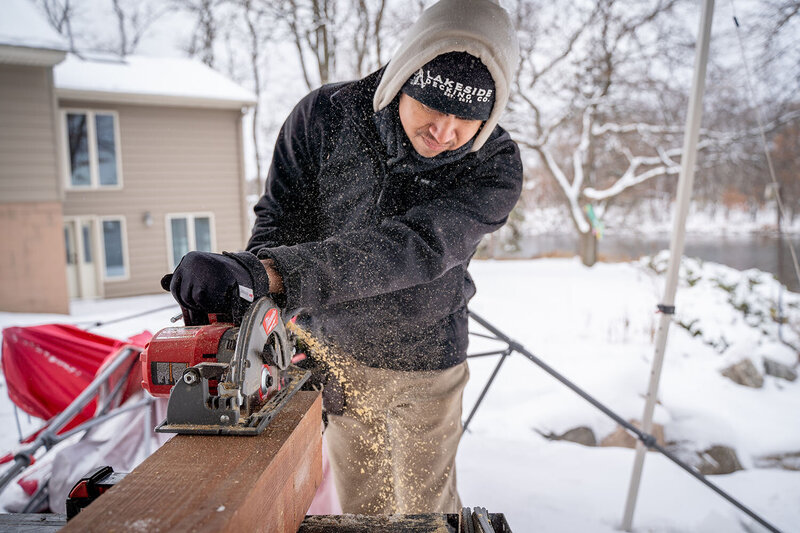 A man wearing cold weather gear on a snowy Minnesota day, sawing a large piece of wood with a circular saw and sawdust is flying out the side. 